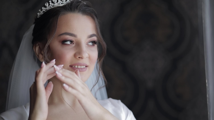 Bride in white boudoir dress stay near window. Wedding morning preparations. Luxury bride with fashion hairstyle and makeup in a hotel or apartment. Beautiful, lovely woman in night gown and veil | Shutterstock HD Video #1062803353