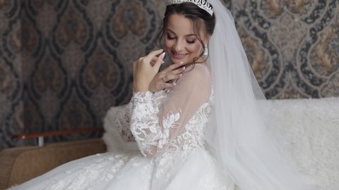 Beautiful, lovely bride in wedding luxury dress and veil. Pretty and well-groomed gorgeous woman sitting on the bed. Wedding preparation, happy moments, couple goals, morning of a bride. Slow motion