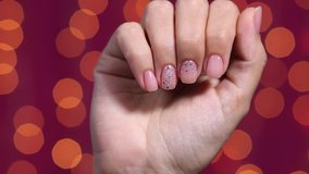 Closeup view video of beautiful manicured female hands isolated on bright red bokeh holiday background. Fingernails painted in pastel natural pink color, two nails decorated with silver shiny glitter.