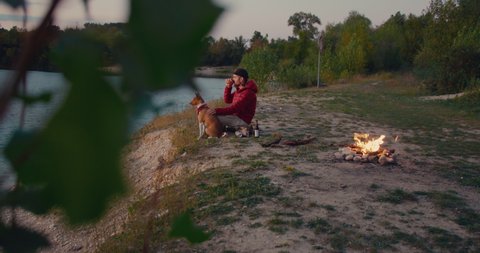 Man and his best friend, brown basenji puppy dog sit on sea or lake shore, watch sunset next to campfire on weekend road trip. Traveler tourist or active lifestyle outdoors lover enjoy nature at camp Video de stock