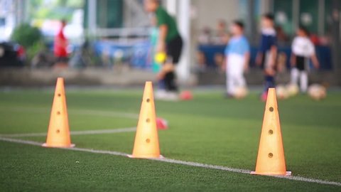 Selective focus to orange marker cone is soccer training equipment on green artificial turf with blurry coach and kid soccer players training background. Blurry Sport background.