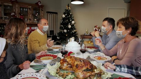 Happy family members wearing surgical masks at the Christmas Thanksgiving table at home in the year of the Covid-19 Pandemic. Grandfather proposing a toast. New rules, new health regulations.