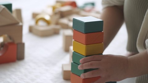Close-up of child's hand playing with wooden blocks on the white floor mat at home. 2 years old little toddler baby girl. Collapses when trying to stack high. Messy building blocks background