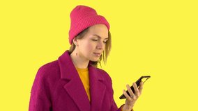 A Flirtatious Young Cheerful Woman in a Red Coat and Hat Uses a Mobile Phone, Typing a Message. A Beautiful Caucasian Woman Taps the Screen of her Smartphone and smiles, Yellow Background.
