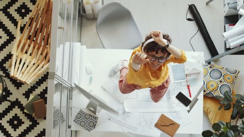 Top down shot of young female architect in wireless headphones dancing to favorite music on office desk, smiling and looking up at camera