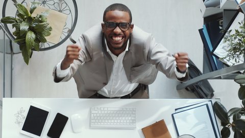 Top down shot of young Afro-American businessman in formalwear typing on computer at office desk, then looking up, raising arms and yelling in excitement at camera while celebrating success