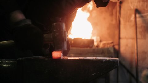 Blacksmith hammering red hot metal piece on an anvil. The hammer of smith beat on glowing hot metal and sparks fly in all directions. Handmade Blacksmithing in a Workshop.