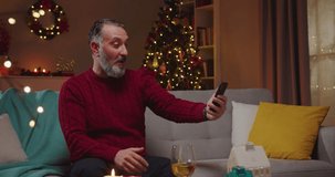 Portrait of cheerful Caucasian handsome adult grey-haired man speaking on video call online on smartphone while sitting on couch in decorated room in evening with glowing xmas tree on background