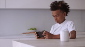  teenager boy playing games on phone. addicted child sit at modern kitchen and gaming online, using smartphone indoors. technology and device concept