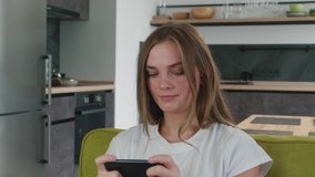Gambling young woman in white t-shirt play mobile game on cell phone at home. Attractive smiling female enjoying smartphone video games sitting on sofa at living room.