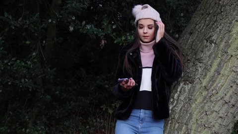 Young attractive woman in a park leaning against a tree in the winter time, looking at her phone online dating swiping left and right on a dating app, liking some of the profiles and not liking some