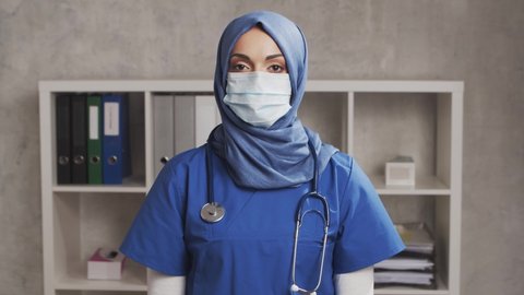 Professional Muslim medical doctor works in hospital office. Young and attractive Middle Eastern female physician in protective mask. Medical concept.