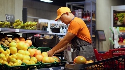 Chinese retail store worker in orange uniform arranging fresh fruits in supermarket section indoors. Asian ethnicity. People and profession. Grocery shop. Consumerism.