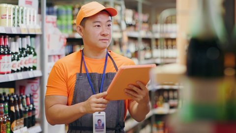 Chinese workman seller employee wearing uniform counting bottles in alcoholic section managing number and quality measures. Supermarket. Profession concept.
