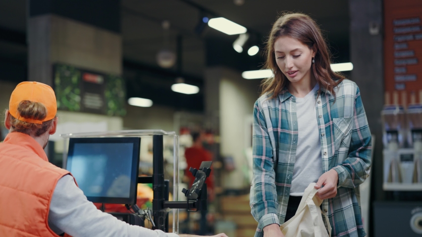 Young beautiful woman standing at cash register communicating with cashier packing her products in plastic bags. Supermarket. Grocery store shopping. Royalty-Free Stock Footage #1062834613