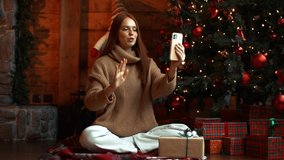 Charming smiling young woman showing festive interior, Christmas tree and xmas gift box during video call on mobile phone at eve of winter holidays sitting on floor at cozy dark living room.