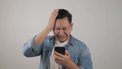 Attractive young Asian man reading texting chatting  on his phone, bad news, sad crying expression, over grey