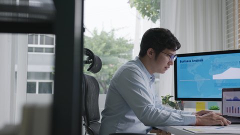 Side view of young attractive asian thai man busy work multiple screen computer or smart tablet on table desk at home in freelance data analyst, data science scientist for business.