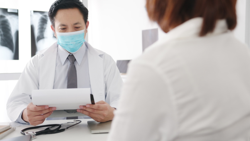 Serious Asia male doctor wear protective mask using clipboard is delivering great news talk discuss results or symptoms with female patient in hospital office. Lifestyle new normal after corona virus. Royalty-Free Stock Footage #1062848242