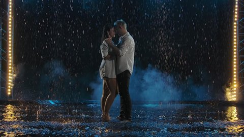 A passionate young couple in white stands in the rain with their bodies pressed together and tenderly caressing each other. Slow motion.