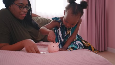 African American mother teach her daughter or little girl to collect money by put in piggy bank and stay on bed in bedroom. They look happy and enjoy together of good bonding family.