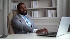African-American bearded man in a gray suit and shirt. The businessman is in a bright office at the workplace, in front of him on the table is a laptop. He talks via video link and smiles and laughs.