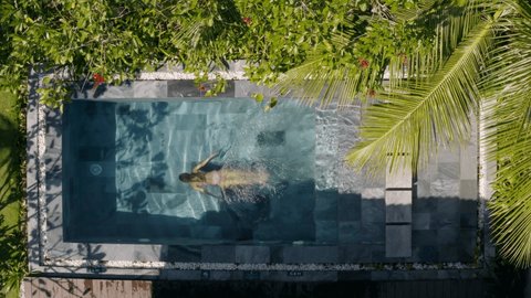 Top down view drone shot: Woman swimming in private swimming pool. Aerial drone shot of woman swimming and splashing inside private villa pool.