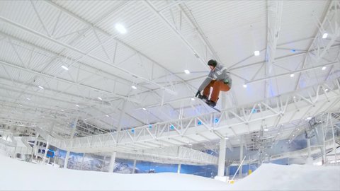 Expert snowboarder at an indoor slope does a jump trick and grabs the snowboard in air - follow shot