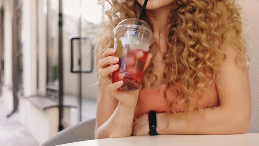 Young beautiful smiling hipster woman with afro curls hairstyle. Woman in summer trendy clothes sitting in veranda cafe.Model drinking fresh cocktail smoothie drink in plastic cup with straw