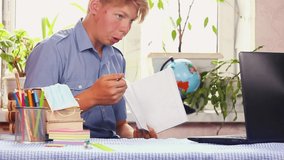 a young white teenager in a blue shirt sits at his desk in his room and shows homework from a notebook to the teacher through a laptop. distance learning during quarantine