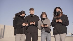 Group of friends with face masks watching a funny video on a smartphone. Millennials laughing and having fun together outdoors. New normal lifestyle in the covid-19 season.