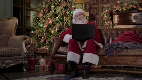 Santa Claus communicates on laptop via video communication with children, sings Christmas songs and gestures with his hands while sitting on the couch. Santa communicates remotely to pandemic covid19