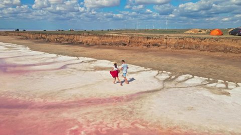 drone flying around lovely young couple running together hand to hand along red clay shore of beautiful pink lake with salty coast. lovebirds tourists walking on white salty lake coast.