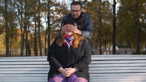 Young man covering eyes and surprising obese girlfriend sitting and waiting on bench in autumn park. Happy loving couple meeting outdoors having romantic date