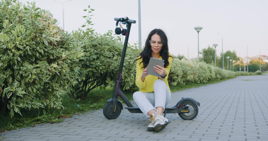 Good-looking stylish thoughtful young woman in yellow sweater and light blue jeans sitting on e-scooter and use tablet pc near decorative bushes
