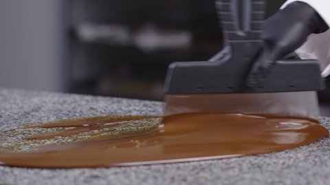 confectioner pours melted milk chocolate on granite table for tempering. Chocolatier stirring with spatula tempered liquid chocolate side view. Cook tempers chocolate. World Chocolate Day concept
