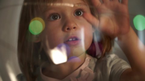 Close up of little child rotates transparent ball of blurred multicolored glowing lights. Girl looks at camera through magic sparkling colored lights. Christmas and New year concept.