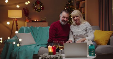 Caucasian joyful adult married couple in festive holiday mood at home speaking on video call with kids on laptop on New Year sitting on sofa. Christmas glowing tree concept. Winter holidays स्टॉक व्हिडिओ