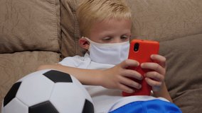 A little boy in a medical mask is lying on the couch at home with a soccer ball and playing games on his phone. Quarantine and distance learning due to the coronavirus outbreak.