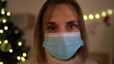 Close up Portrait of lonely woman wearing medical mask with sad emotion. Against the background of a Christmas tree. Coronavirus pandemic. Christmas and covid-19