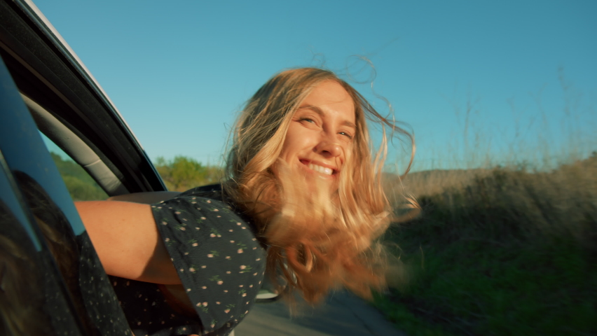 Portrait of happy young woman hang out of car window on sunny road trip day, enjoy views and fresh air. Travel blogger or social media influencer enjoy travelling on warm day