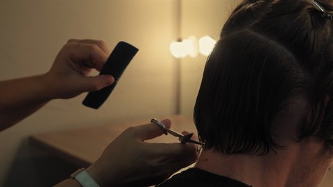 Back view hairdresser woman parts in sections mans head to trims back of his head pulling his hair cuts off the ends in semi lit light room. High quality 4k resolution footage. 