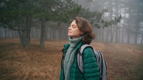 A cinematic shot of a young cheerful woman makes a deep breath and enjoy the fresh air while walking in the misty forest on a fall day.