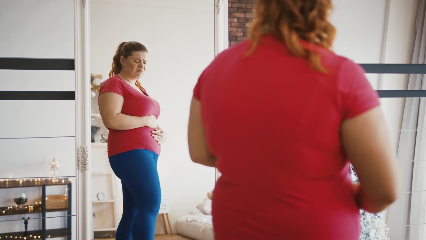 Obesity problem. Young overweight woman feeling sad because of excess weight, looking at herself at mirror Royalty-Free Stock Footage #1062892486
