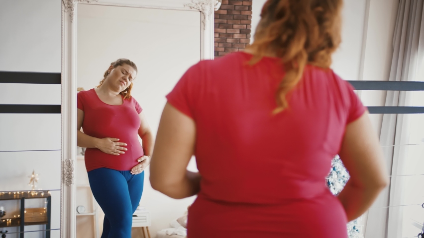 Obesity problem. Young overweight woman feeling sad because of excess weight, looking at herself at mirror Royalty-Free Stock Footage #1062892486