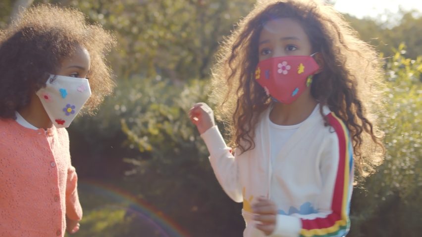 Happy mixed race little girls in safety mask hugging and smiling in park. Portrait of adorable african preschool sisters wearing protective mask embracing outdoors