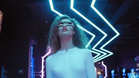 Fashion young woman in futuristic glasses and white costume walks in night club in city of future. Blue and violet neon lights. Trendy stylish millennial female. VR club, future technology. Following