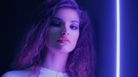 Close-up portrait of beautiful trendy woman wearing white outfit posing and looking into camera standing by illuminated wall in night club. Futuristic style, sensual look clubber in neon violet light