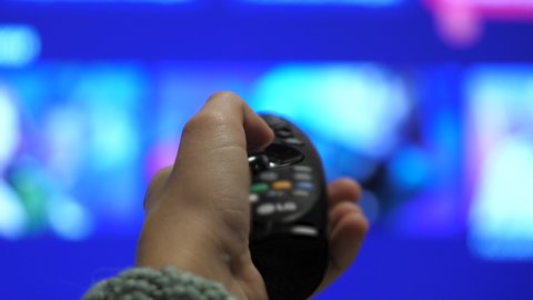 Man's hand selects internet tv channels with remote control, close-up. Person controls TV using a modern remote control. A man watches smart TV and uses black remote control. Blurry tv scrolls pages
