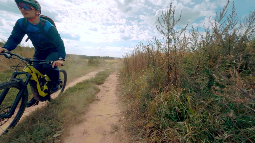 Professional sportsman biker extreme rides fast modern mtb e-bike bicycle  along rural ground road among autumn field past forest and bushes under  cloudy sky. cinematic shot by sport fpv drone.